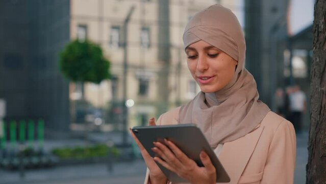 Happy arabian business woman wearing hijab sitting near office centre, working outdoors, using tablet for work matters. Female manager working on tablet on the business centre background