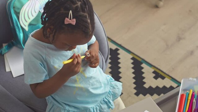 From above shot of playful African American toddler girl drawing with colorful crayons on her light blue t shirt while playing alone at home