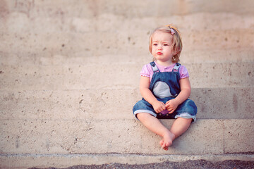 Fototapeta premium A cute little girl is sitting on the steps of the pier on the seashore. Street portrait of a little girl on a walk. Young blonde in the rays of the setting sun