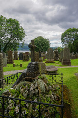 Fototapeta na wymiar Luss, Scotland: A grave overgrown with mosses and topped with a stone cross in the 1875 Luss Parish Church cemetery on a cloudy day, with Loch Lomond in the distance.