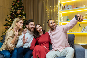 New Year's party home diverse group of friends, men and women, sitting on the couch, taking a selfie together, using a smartphone, guests looking at the phone, talking on a video call for Christmas