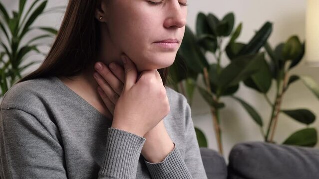 Symptoms from flu season in winter concept. Close-up of unhealthy young caucasian woman  sitting on couch at home touching her neck and suffering from sore throat, angina, hard to swallow, voice loss