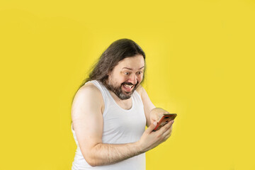 A joyful unshaven man in a white undershirt pokes his finger at the screen of a mobile phone with a...