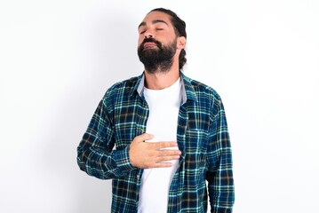 Satisfied smiling young bearded hispanic man wearing plaid shirt, keeps hands on belly, being in good mood after eating delicious supper, demonstrates she is full. Pleasant feeling in stomach.