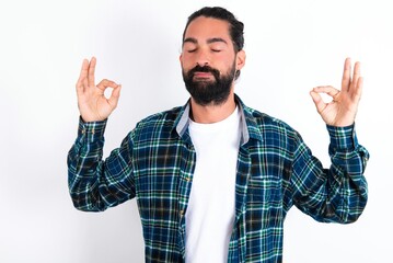 Fototapeta premium young bearded hispanic man wearing plaid shirt over white background doing yoga, keeping eyes closed, holding fingers in mudra gesture. Meditation, religion and spiritual practices.