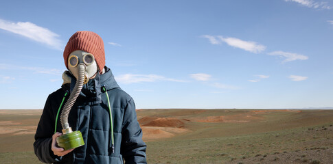 Portrait of a boy in a gas mask and warm clothes against the backdrop of the desert on a sunny day. Dramatic landscape with cloudy sky without people and plants. Apocalypse concept. Copy space - obrazy, fototapety, plakaty