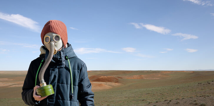 Portrait Of A Boy In A Gas Mask And Warm Clothes Against The Backdrop Of The Desert On A Sunny Day. Dramatic Landscape With Cloudy Sky Without People And Plants. Apocalypse Concept. Copy Space