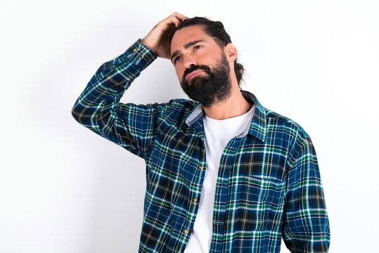 young bearded hispanic man wearing plaid shirt over white background saying: Oops, what did I do? Holding hand on head with frightened and regret expression.