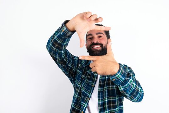 Positive young bearded hispanic man wearing plaid shirt over white background with cheerful expression, has good mood, gestures finger frame actively at camera.