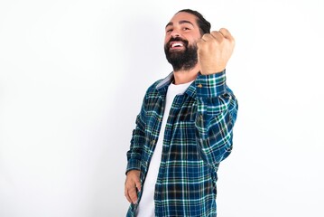 Fototapeta premium Overjoyed young bearded hispanic man wearing plaid shirt over white background glad to receive good news, clenching fist and making winning gesture.