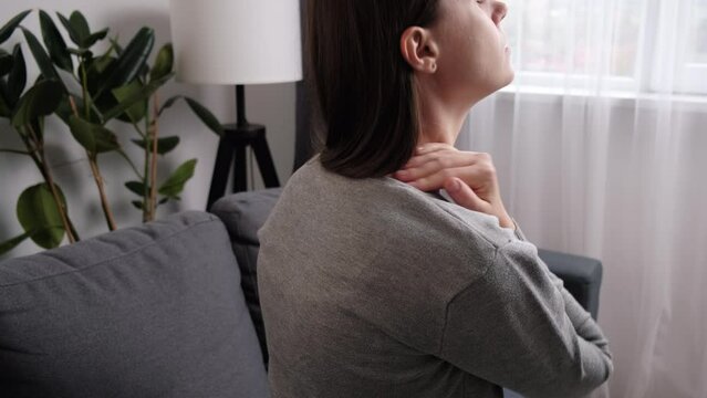 Tired young brunette woman sitting on sofa at home massaging stiff sore neck tensed muscles reduces painful feelings. Shoulder back pain fibromyalgia osteoarthritis senile chronic diseases concept