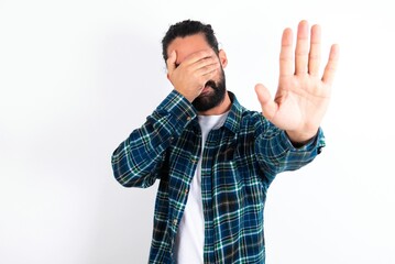 young bearded hispanic man wearing plaid shirt over white background covers eyes with palm and...