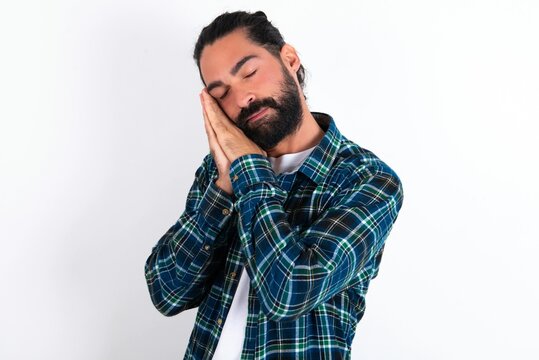 Relax and sleep time. Tired young bearded hispanic man wearing plaid shirt over white background with closed eyes leaning on palms making sleeping gesture.