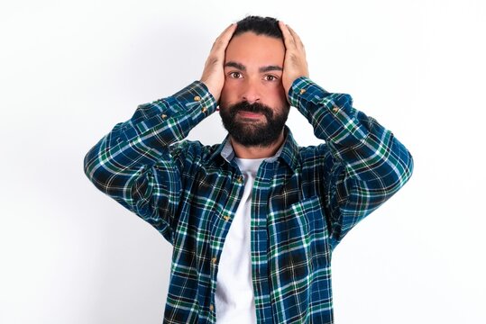 Frustrated young bearded hispanic man wearing plaid shirt over white background plugging ears with hands does not wanting to listen hard rock, noise or loud music.