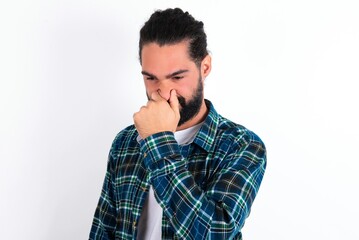 young bearded hispanic man wearing plaid shirt over white background, holding his nose because of a bad smell.