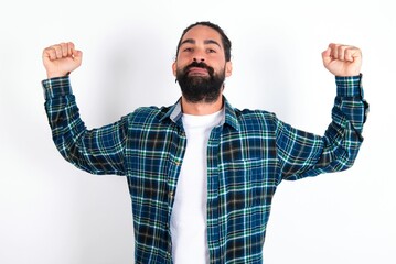 Waist up shot of young bearded hispanic man wearing plaid shirt raises arms to show muscles feels confident in victory, looks strong and independent, smiles positively at camera. Sport concept.