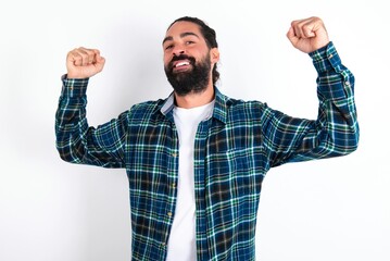Naklejka premium Yes I am winner. Portrait of charming delighted and excited young bearded hispanic man wearing plaid shirt raising up fist in triumph and victory smiling achieving success grinning from delight.