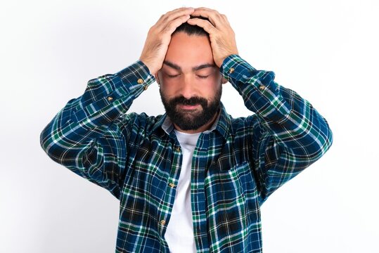 young bearded hispanic man wearing plaid shirt over white background holding head with hands, suffering from severe headache, pressing fingers to temples