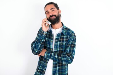 Portrait of a smiling young bearded hispanic man wearing plaid shirt over white background talking on mobile phone. Business, confidence and communication concept.