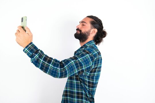Portrait of a young bearded hispanic man wearing plaid shirt over white background  taking a selfie to send it to friends and followers or post it on his social media.
