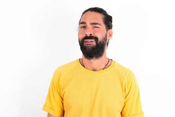 Portrait of dissatisfied young bearded hispanic man wearing yellow T-shirt over white background smirks face, purses lips and looks with annoyance at camera, discontent hearing something unpleasant