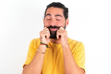 Pleased young bearded hispanic man wearing yellow T-shirt over white background with closed eyes keeps hands near cheeks and smiles tenderly imagines something very pleasant