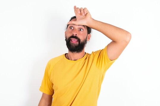 young bearded hispanic man wearing yellow T-shirt over white background gestures with finger on forehead makes loser gesture makes fun of people shows tongue