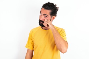 Disappointed dejected young bearded hispanic man wearing yellow T-shirt over white background wipes tears stands stressed with gloomy expression. Negative emotion