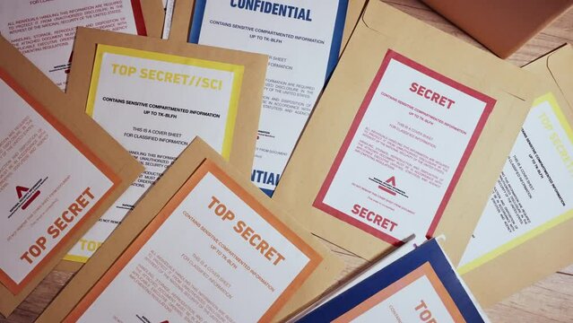 Stack of various yellow manila folders with top secret and classified labels. Confidential files and documents inside envelopes stacked on desk or floor. Leaked or uncovered by agency or police force
