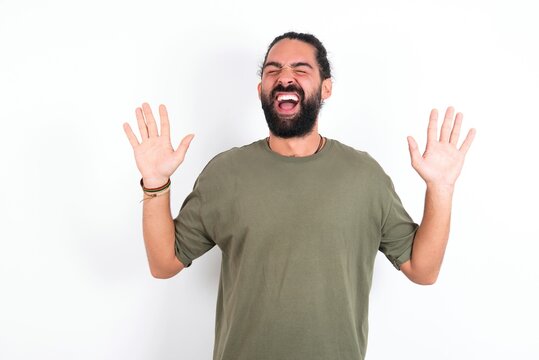 Emotive young bearded hispanic man wearing green T-shirt over white background laughs loudly, hears funny joke or story, raises palms with satisfaction, being overjoyed amused by friend