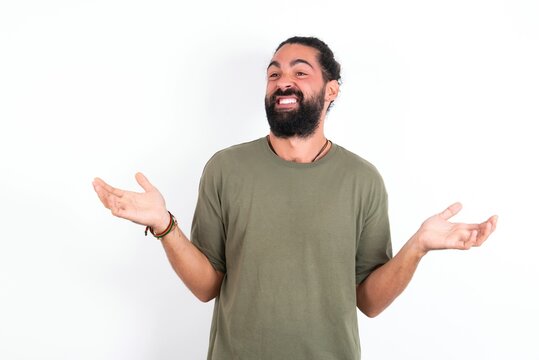 Clueless young bearded hispanic man wearing green T-shirt over white background shrugs shoulders with hesitation, faces doubtful situation, spreads palms, Hard decision