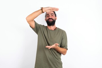 young bearded hispanic man wearing green T-shirt over white background gesturing with hands showing big and large size sign, measure symbol. Smiling looking at the camera.