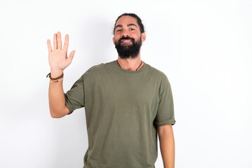 young bearded hispanic man wearing green T-shirt over white background Waiving saying hello happy and smiling, friendly welcome gesture.