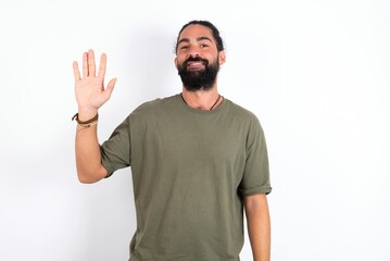 Fototapeta premium young bearded hispanic man wearing green T-shirt over white background Waiving saying hello happy and smiling, friendly welcome gesture.