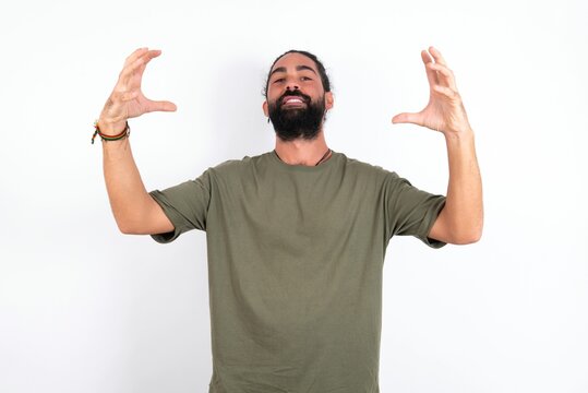 young bearded hispanic man wearing green T-shirt over white background Shouting frustrated with rage, hands trying to strangle, yelling mad.