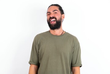 young bearded hispanic man wearing green T-shirt over white background winking looking at the camera with sexy expression, cheerful and happy face.