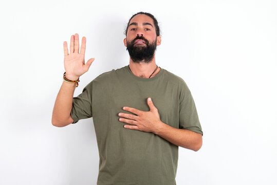 young bearded hispanic man wearing green T-shirt over white background Swearing with hand on chest and open palm, making a loyalty promise oath
