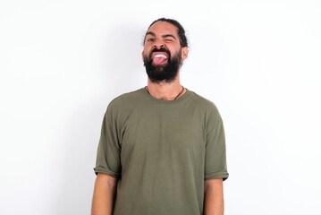 Fototapeta premium young bearded hispanic man wearing green t-shirt over white background sticking tongue out happy with funny expression. Emotion concept.
