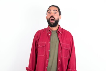 Obraz premium Oh my God. Surprised Caucasian man with beard wearing red shirt over white background stares at camera with shocked expression exclaims with unexpectedness,