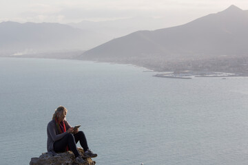 Mature woman sits on rock above sea and mountains and texts