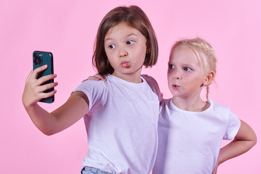 Two cute little girls taking a selfie with a phone, using a front camera; pink studio background
