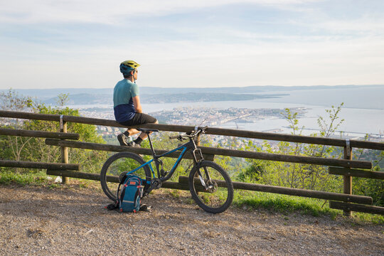 Mountain biker sitting on wooden railing and looking at sea and city