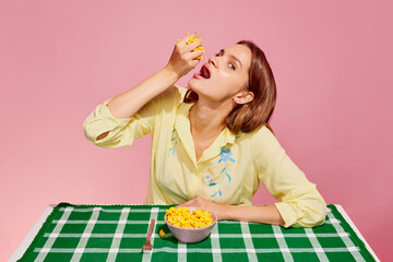 Healthy eating. Food pop art photography. Young girl tasting sweet canned corn isolated over pink background. Concept of food, creativity.