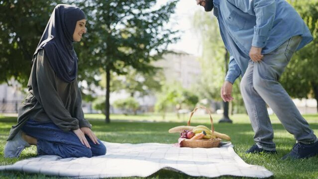 Happy muslim couple laying blanket on grass in park, picnic, outdoor activities