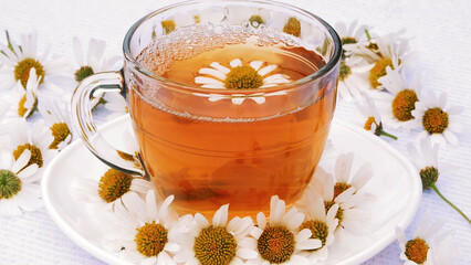 Close-up, transparent, glass cup with chamomile tea, on a white saucer. Everything is decorated with chamomile flowers. High quality photo