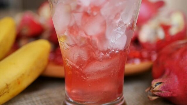 pour a red refreshing drink into a glass with ice cubes close-up on the background of many ripe pomegranates and bananas. Refreshing cocktail with pomegranate red grist structure
