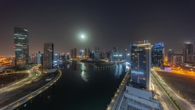 Cityscape of skyscrapers in Dubai Business Bay with water canal aerial during night timelapse with Moon setting down. Modern skyline with illuminated towers and lights turning off