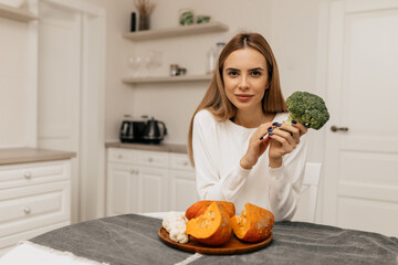 Portrait of pretty woman holding green vegetables at home kitchen. Pretty housewife tasting fresh meal at home. Cheerful female chef enjoying food taste indoors. Joyful woman face at modern kitchen.