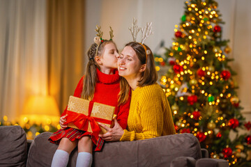 Obraz na płótnie Canvas loving daughter kisses mom at home, sitting on couch against background of a Christmas tree. child and woman laughing happily, holding gift box in their hands Give each other gifts for Christmas