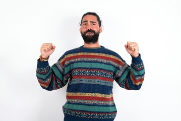 Fototapeta premium Irritated Caucasian man with beard wearing sweater over white background blows cheeks with anger and raises clenched fists expresses rage and aggressive emotions. Furious model